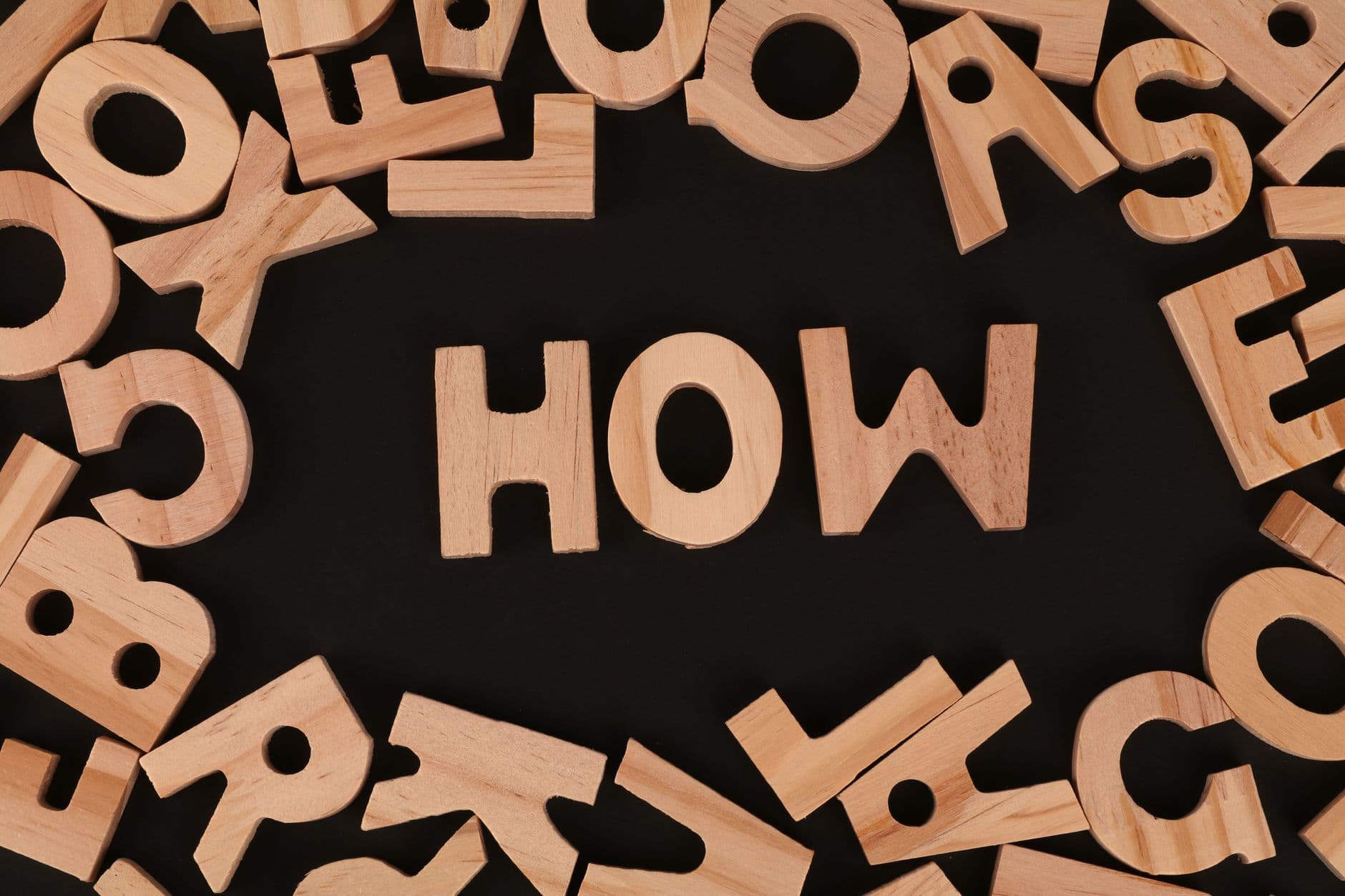 Top view of scattered wooden letters with 'HOW' centered on a dark surface, promoting inquiry.